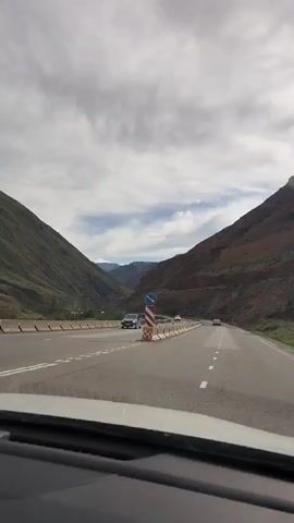 This video shows a view from the inside of a car driving on a paved road that curves to the right. The road is bordered by concrete barriers and has white dashed lines marking the lanes. On the left side of the road, there are steep green hills, and on the right side, there are steep reddish-brown hills. Several cars are visible on the road ahead, and the sky is cloudy.