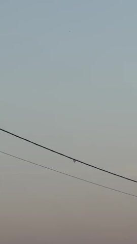 A hot air balloon is visible in the sky, seemingly suspended over power lines. The sky is clear and hazy, suggesting it may be early morning or late evening. The balloon is small in the frame, indicating it is likely at a significant distance. There is a roof of a building visible at the bottom of the frame.