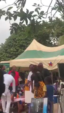 The video shows a group of people gathered in what seems like an outdoor event or party.  They are standing near a tent-like structure with green and white stripes.  A child in a yellow shirt stands in the foreground.  Some people are dancing, while others are talking and interacting. There are several adults of both sexes.  The overall atmosphere appears lively and festive.