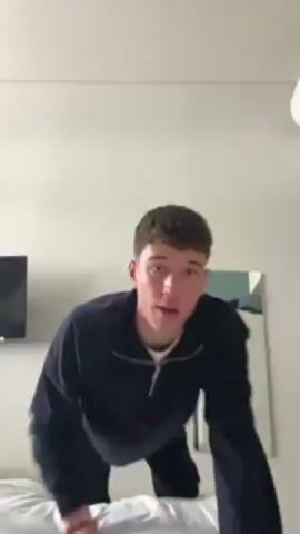 A young man in a dark blue, zip-up sweater and dark pants is standing on a bed. He is breathing heavily and looks straight ahead. Behind him is a white wall, a television on a stand, and a white cabinet with a mirror on the door. The room is well-lit. The man appears to have just finished some sort of physical activity.