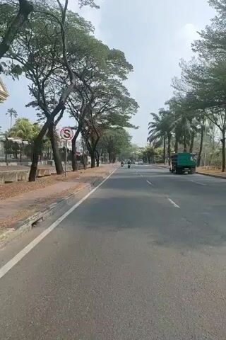 The video shows a street view from a moving vehicle. The road is paved and has lane markings. On the left side of the road, there is a sidewalk, a traffic sign with a red circle and a diagonal line, trees, and some buildings. On the right side, there are more trees and a green truck. Several motorcycles are visible on the road ahead. The sky is bright and cloudy. The overall environment appears to be an urban or suburban area with greenery.