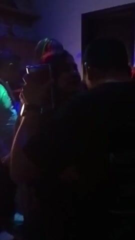 In a dimly lit room with a blue and green strobe light effect, a group of people appear to be gathered, possibly at a party or small gathering. A man wearing a white t-shirt and a lanyard is visible on the left. To the right, a person is holding up a phone to record something, their face obscured by their dark hair. In front of them, a woman with curly hair is seen smiling. An air conditioning unit is mounted on the wall near a dark doorway. The camera pans slightly up to reveal a shelf with various items on it.