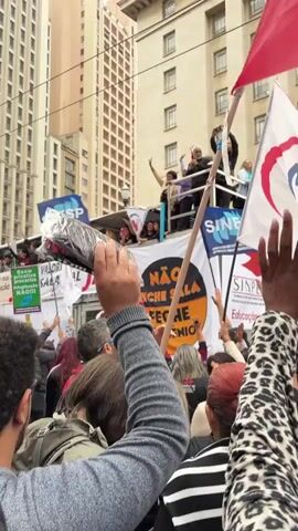 The video shows a crowd of people protesting in a city street. People hold signs and flags, with the words "SINESP" and "NAO FECHE SALAS" visible. There are tall buildings in the background. Some people stand on top of a truck waving and raising their arms. The protesters are also raising their arms. The day is overcast.