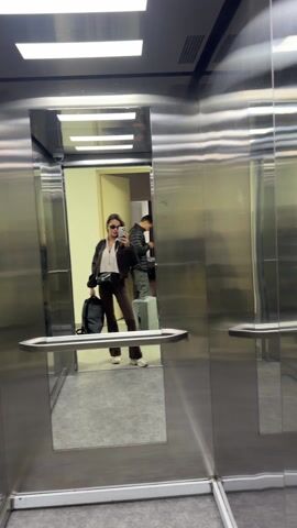 In an elevator, a young woman stands near the door, taking a selfie in the mirrored wall. She wears sunglasses, a brown jacket, a light pink top, brown pants, and white sneakers. She has a black bag slung across her body and holds a gray backpack. Behind her, near the back wall, a man stands with a light blue suitcase and appears to be looking at his phone. The elevator has a stainless steel interior with a horizontal handrail. The doors are closing.