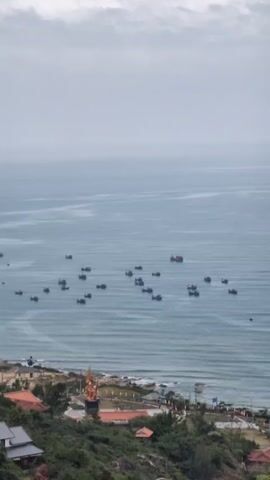 The video shows a coastline with many small fishing boats on the water. The sky is overcast.  The camera pans to reveal a golden statue near a red building and a pagoda on a nearby hill.  The landscape is lush and green.