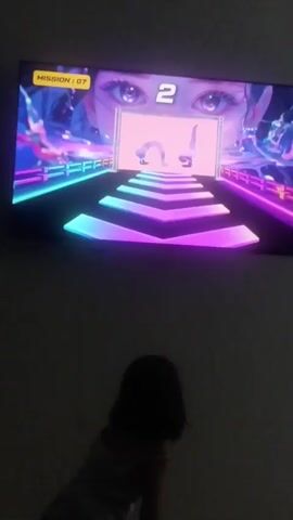 A young child, facing away from the camera, stands in front of a large television screen. The screen displays a video game with bright, neon-colored graphics. The game appears to be a rhythm or exercise game, as the screen shows a large anime-style character's face with glowing eyes, and below it, a neon pathway leading to a central screen. This central screen shows two figures in yoga poses, a bridge pose and a handstand pose, with the number "2" prominently displayed. The child is looking at the television and appears to be mimicking the poses shown on the screen, raising their arms in a gesture that could be related to the game's prompts. The environment is dimly lit, suggesting the video is being watched in a darkened room. There are no other people visible in the frame, and the focus is on the interaction between the child and the game.
