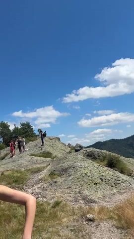 A group of people are hiking on a rocky mountain terrain with dry grass and some trees. The sky is clear and blue with some white clouds. In the foreground, a person is holding a black piece of cloth and looking at the camera. In the background, there are several other hikers. The video pans across the landscape, showing the vast mountainous scenery.