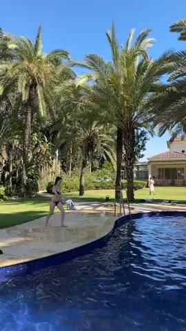 A young woman in a black bikini runs and jumps into a blue swimming pool surrounded by palm trees. She splashes into the water and surfaces with her hands to her head. In the background, a large house and a lawn are visible, with several people gathered around a table. The sky is clear and blue.