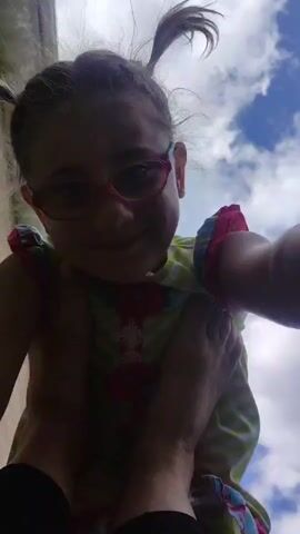 A child with glasses and pigtails is held upside down. The child wears a colorful dress. The child is looking up towards the camera and smiling. There are some clouds in the background.