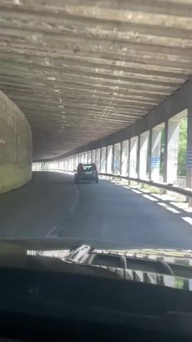 The video shows a rear-facing view from inside a vehicle as it travels through a tunnel. A dark-colored SUV is visible ahead on the same road. The tunnel has concrete walls and a concrete ceiling with visible horizontal supports. Sunlight streams in from openings on the right side of the tunnel, illuminating the road and creating shadows. The road appears to be asphalt and has a white lane divider. The overall impression is of a clear day outside the tunnel.