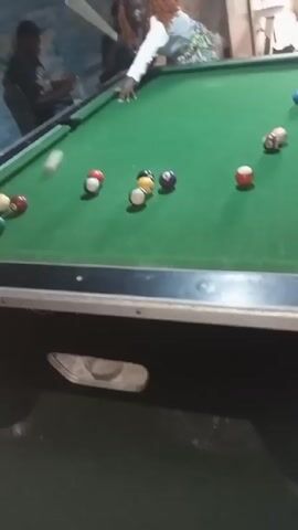 A medium shot shows a pool table with various pool balls arranged on its green felt surface. A person in a white top is poised to make a shot. The camera shakes slightly after the shot. Two additional people are partially visible in the background. The pool table is black with silver trim. The room appears to be dimly lit with a rough wall texture in the background.