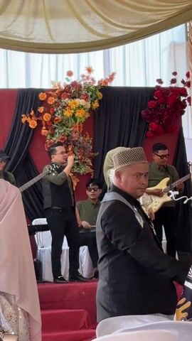 The video shows a musical ensemble performing on stage at what appears to be a wedding. Two singers, a man and a woman, are center stage, each with a microphone. The woman is wearing a hijab and a sparkly top with a long green skirt. The man is wearing glasses, a dark vest over a green shirt, and black pants. Behind them are three musicians: a guitarist, a bassist, and a drummer seated at a small electronic drum set. The musicians are all dressed in similar shades of green. The stage backdrop is red with black curtains draped on either side, and adorned with floral arrangements in orange, yellow, and red. Guests are visible in the foreground, with one woman in a light blue dress walking across the frame. The atmosphere suggests a celebratory and musical occasion.