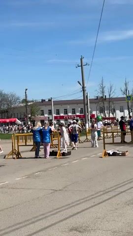 The video shows a street scene on a bright, sunny day, with a clear blue sky. There's a road with yellow metal barriers, beyond which a crowd of people are gathered. People walk across the road in various directions. A statue painted gold sits near the center of the frame. A building with signage is visible in the background. Overhead power lines cut across the sky.