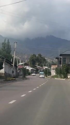 The video shows a street in a village with mountains in the background. The road is paved and has white dashed lines. There are several houses on the side of the road, as well as trees and utility poles. Three white cars are driving on the road. The sky is cloudy. The video appears to be shot from a moving vehicle.