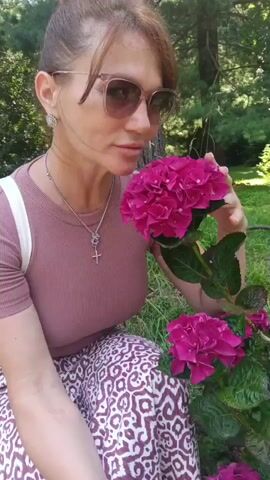 A woman with brown hair pulled into a bun and wearing sunglasses, a mauve t-shirt, and a patterned skirt is squatting in front of a hydrangea bush. She is holding a bloom of pink hydrangeas to her nose and smelling them. She then looks up and smiles at the camera. The background is a wooded area with green foliage and trees.