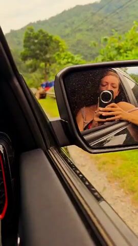 The video shows a car driving along a winding road through a lush, green mountainous area. In the side mirror of the car, a woman with curly hair is visible, holding a smartphone and appearing to be recording herself. Other vehicles, including a yellow construction vehicle, can be seen on the road. The road has a guardrail along the edge in some places, and the landscape is dominated by dense vegetation.
