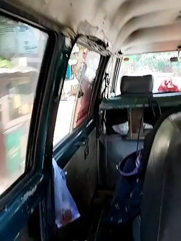 The video shows the interior of a vehicle, likely a van or minibus, from a first-person perspective, suggesting the viewer is a passenger. The camera is angled towards the rear of the vehicle. The interior appears worn and somewhat dated. The headliner shows signs of peeling and damage, particularly near the edges. The windows are visible, with the left side showing a blurred view of the outside environment, including some structures and trees. The right side windows offer a clearer view of the passing scenery.

There are seats visible, upholstered in a dark material, showing signs of wear. In the foreground, a dark-colored backpack or bag with purple accents rests on a seat. Further back, a person wearing a dark headscarf or hijab is seated, their back to the camera. The headscarf covers their hair and neck, and they appear to be looking forward or slightly to the side. The overall lighting suggests daytime, possibly with sunlight filtering into the vehicle. The environment outside the vehicle appears to be an urban or semi-urban setting with buildings and vegetation. There are no overt actions or interactions depicted, just a static view from within the moving vehicle.