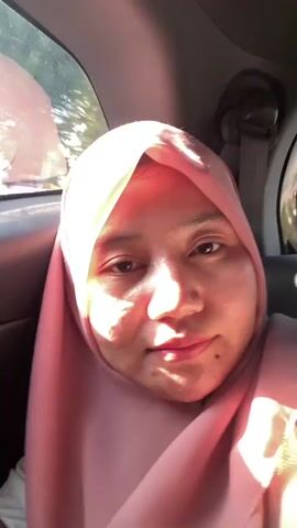 A woman wearing a pink hijab is filmed from the chest up, looking towards the camera with her eyes half-closed. She appears to be in a car, with sunlight streaming in through the windows, creating lens flares. The background outside the windows is blurry, showing trees and buildings. The woman is smiling slightly. The camera is shaky and moves erratically, indicating it is likely handheld and in motion.