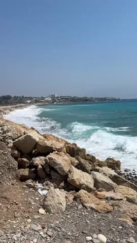 The video shows a rocky coastline with waves crashing against the shore. The water is a vibrant turquoise color, and the sky is a clear blue. In the distance, there are buildings that appear to be part of a town or resort. The foreground of the video is dominated by large rocks and pebbles, with some scattered debris. The waves are moderate in size, creating white foam as they break. The overall impression is of a sunny day at the beach, with the natural beauty of the ocean and coastline on display.
