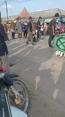 A group of people are walking down a street. The person in the foreground on the left is wearing a black shirt and tan pants and is partially cut off by the frame. To their right, a motorcycle is parked, with its front wheel and chrome spokes visible. In the middle ground, a group of people are walking and interacting. Some are wearing traditional clothing with brown and white patterns, while others are in dark clothing. One individual is wearing camouflage pants and a black t-shirt, holding what appears to be a camera. Another person is crouching down. In the background, there are buildings with awnings and signs, and more people are visible. The street is paved with asphalt and appears to be a public area, possibly a market or a street fair. The lighting suggests it is daytime, with shadows cast on the ground.