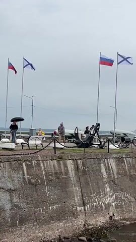 The video shows a section of a concrete embankment along a body of water. Several people are visible on the embankment, some holding umbrellas, suggesting overcast weather. There is a chain fence separating the embankment from an area where various military objects are displayed, including what appear to be missiles, torpedoes, and possibly aircraft components. Russian flags and naval flags are flying from poles. In the background, buildings and a ship with radar equipment are visible. The water in the foreground is dark and appears choppy.