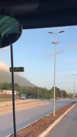 The video shows an airplane descending from the sky, viewed from inside a vehicle. The vehicle appears to be stationary on a road undergoing construction. A mountain is visible in the background, partially obscured by clouds. A few streetlights stand along the side of the road. The sky is mostly clear with a few sparse clouds.
