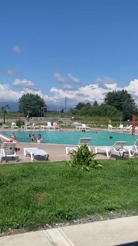 A wide shot of an outdoor swimming pool on a sunny day with scattered clouds. People are swimming in the turquoise water and lounging on white deck chairs around the pool. In the background, there are trees, a stone structure with water features, and a power pole. The pool area is surrounded by green grass and a wooden deck.