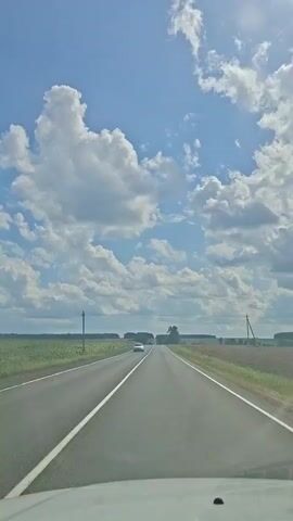 The video shows a view from inside a vehicle driving on a paved road. The road has a white line marking the lane. On either side of the road are fields of green crops. The sky is blue with large, white, fluffy clouds. In the distance, there are a few trees and power lines. Another vehicle, a white car, is visible in the distance driving in the same direction as the camera vehicle.