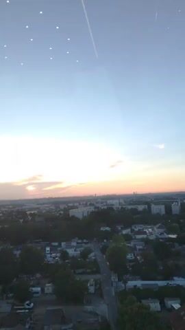 The video shows a panoramic view of a city skyline during sunset. The sky is a gradient of blue, transitioning to orange and pink hues as the sun sets. Clouds are scattered across the sky, catching the light and adding to the vibrant colors. In the foreground, there are trees and buildings, suggesting a residential area. The middle ground reveals more buildings, including apartment complexes. In the distance, a denser urban landscape with taller buildings, including some skyscrapers, can be seen. The lighting suggests it is either sunrise or sunset. There are no people or active interactions visible in the video. The camera is stationary, offering a static shot of the cityscape.