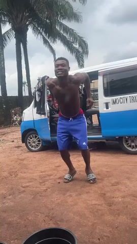 A young Black man with a muscular build is shown jumping in the air. He is shirtless and wearing blue athletic shorts with white stripes down the sides and red trim around the waist. He is also wearing black sandals. He is smiling and appears to be happy. Behind him is a white and blue van with the words "IMO CITY TRA" visible on the side. There are palm trees in the background and the ground is covered in reddish-brown dirt. The sky is overcast. The man is in the center of the frame and appears to be the focus of the video.