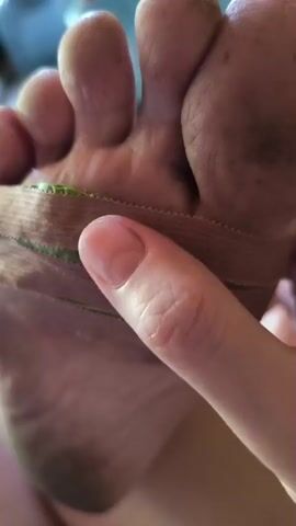 The video shows a close-up view of a person's foot. A finger is touching a band of tan tape wrapped around the arch of the foot. The tape has two thin green lines running through its center. The toes of the foot are visible, and the sole of the foot appears dirty. The background is out of focus but suggests an indoor environment with furniture.
