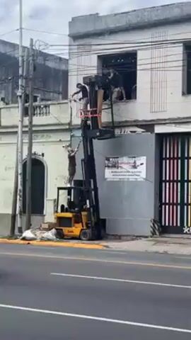 The video shows a street scene where several individuals are working on the upper level of a two-story building, accessed by a forklift modified for lifting people. Three individuals are visible in the window of the upper level, while one person stands on the modified platform of the forklift, and another on a small scaffold near the ground. The forklift is positioned close to a partially open garage door. A poster is attached to the structure, though the text is too small to read. In the foreground, various cars drive by on the street, including a black and yellow taxi and other passenger vehicles. Overhead, power lines run parallel to the building. The sky is overcast.