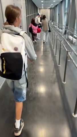 A group of people are walking down a long, enclosed jet bridge or walkway. In the foreground, a person with a ponytail, wearing a gray hoodie, denim shorts, white socks, and black and white sneakers, is walking away from the camera with a white, black, and yellow backpack. Behind this person, several other individuals are walking in the same direction. The walkway has large windows on the right side, offering a view of the outside. Metal railings run along the right side of the walkway, guiding the path of the people. The floor is dark gray and appears to be made of a durable material. The lighting in the walkway is bright and artificial.