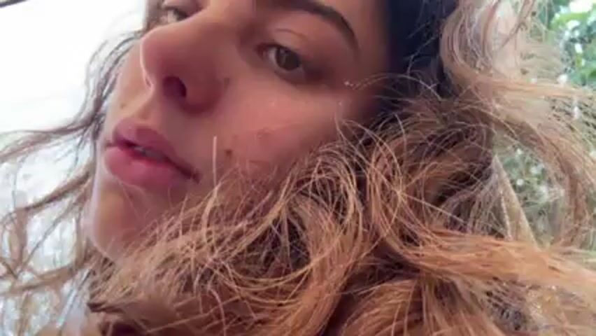 The video shows a close-up of a woman with curly brown hair and light skin. Her hair is somewhat messy and falls around her face. She has neutral expression and looks directly at the camera. There are a few small freckles or blemishes on her face.  The background gradually comes into focus as the camera zooms out.  In the background, there are other people, one man is visible without a shirt on, sitting in a chair.  The environment appears to be outdoors, possibly a rural or wooded area, with some simple buildings and trees visible. The ground looks muddy.