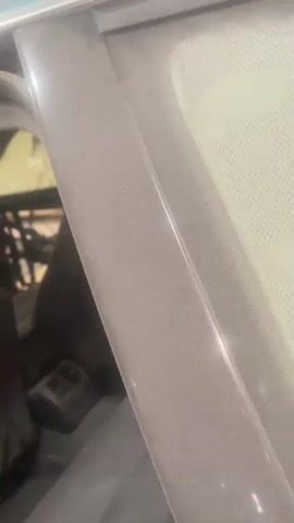 The video shows the interior of a car. The driver's side door is open and the interior panel has been removed, exposing wires and internal mechanisms. The dashboard and steering wheel are visible. The car appears to be in a garage or workshop, with tools and other car parts in the background. The lighting is dim, with overhead fluorescent lights visible.