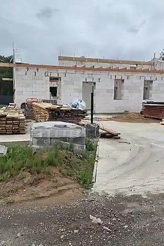 A construction site for a building, likely a house, is shown. The walls are constructed from white cinder blocks. Wooden beams are laid out along the top of the walls, indicating preparation for a roof. Piles of wood planks and pallets are stacked around the construction site, along with a stack of what appears to be more cinder blocks covered in plastic. The ground is a mixture of dirt, gravel, and a concrete or asphalt surface. The sky is overcast and cloudy. The video pans across the scene from left to right. No people are visible.