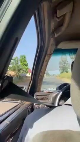 The interior of a car is shown from the passenger seat, focusing on the dashboard, the driver's upper body, and the view out of the front windshield. The driver, an older man with dark skin, is wearing a white shirt and a seatbelt. He is focused on driving down a paved road lined with vegetation and sparse buildings. The dashboard of the car is visibly aged and dusty. The sky outside is bright blue, suggesting it is daytime. The car is moving forward.
