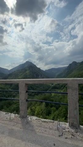 The video shows a scenic view from a bridge under construction. The bridge has a concrete barrier with blue metal railings. The landscape includes lush green mountains and a cloudy sky with the sun peeking through. There are red and white striped warning markers along the bridge. A red truck and some construction scaffolding are visible in the distance. The camera appears to be mounted on a vehicle that is slowly moving along the bridge.