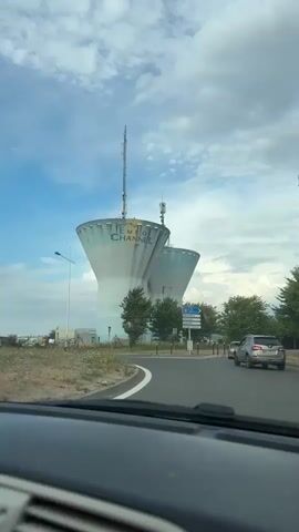 The video shows a view from inside a car. The car is on a road, and the camera is pointing forward. In the distance, there are two large water towers with "EURO CHANNEL" written on them. There are also trees and a blue sky with clouds. A silver SUV is visible on the road in front of the car. There is a road sign with directions to various locations. The car appears to be moving.