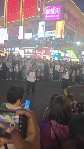 The video captures a lively street performance in what appears to be a bustling city center at night. Two performers, each wearing an LED-lit mask and matching outfits, are dancing for a large crowd of onlookers. The crowd consists of a diverse group of people, including adults and children, many of whom are watching with amusement. Some individuals are recording the performance on their smartphones. The surrounding environment is illuminated by bright neon signs in Chinese characters, and the background buildings are adorned with festive red lanterns. The overall atmosphere is cheerful and engaging, indicative of a community event or street entertainment.