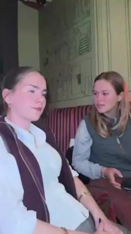 Two young women are sitting next to each other in a booth. The woman on the left is wearing a white collared shirt with a dark burgundy vest, gray pants, and gold hoop earrings. The woman on the right is wearing a gray vest over a long-sleeved shirt and reddish-brown pants. The woman on the left leans her head sideways and closes her eyes, smiling. The woman on the right smiles at the camera. They are sitting against a red-striped cushioned wall. Behind them is a wall decorated with a muted-green drawing of assorted jars and bottles.  A television screen is visible on the left.