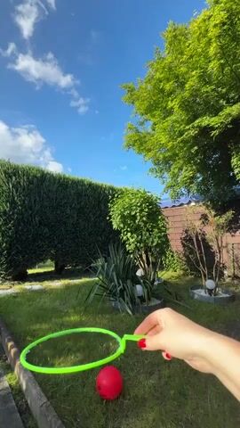 A person holds a green bubble wand over green grass with a red ball. They create a large soap bubble and release it. The bubble floats into the air. The scene is in a backyard with green trees and bushes, a brown wooden fence, a blue sky, and white clouds.