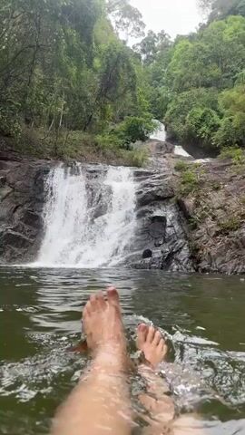 The video shows a pair of feet extending into a body of water, with a waterfall cascading down rocks in the background. The feet appear pale, clean, and relaxed. The water is calm with gentle ripples. The waterfall is surrounded by lush green vegetation, including trees and foliage. The sky is overcast, creating a soft, diffused light. A yellow object floats on the water. The overall scene is tranquil and natural.