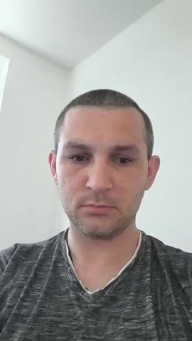 A close-up, vertical, head-and-shoulders shot of a man with short hair and a gray t-shirt. The man is looking directly at the camera with a neutral expression. The background is a plain white wall with a corner visible in the upper left. The lighting appears to be natural, coming from the left. The video is very short, with no discernible action or dialogue.