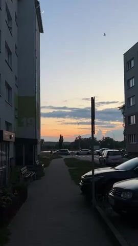The video shows a dusk scene along a sidewalk in an urban environment. Flanked by two gray apartment buildings, the sidewalk is brick-paved. The building on the left has a green and gray stripe with the number 19 prominently displayed. To the right of the sidewalk are several parked cars and a utility pole. In the distance, beyond the parked cars, there's a street with cars driving, a line of trees, and a setting sun, painting the sky with shades of orange and pink. Above the setting sun, clouds are scattered across the sky, with a crescent moon visible high above the buildings.