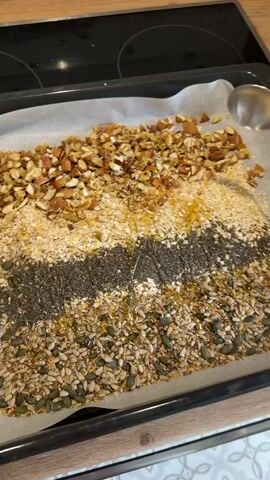 The video shows a close-up overhead view of a baking tray lined with parchment paper. The tray is filled with various ingredients arranged in distinct horizontal rows. From top to bottom, the ingredients appear to be: chopped nuts, followed by a layer of rolled oats and seeds, then a dark layer of chia seeds, and finally a bottom layer of mixed seeds including sunflower and pumpkin seeds. A spoon is actively mixing the ingredients in the oat and seed layer, distributing some of the oil that is present throughout the tray. The background shows a stovetop and a countertop.