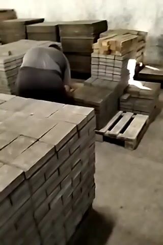 A person is kneeling and working among stacks of grey paving stones in what appears to be a storage or workshop area. The person is wearing a grey shirt and dark pants. The paving stones are stacked in neat piles, some on wooden pallets. In the foreground, a large stack of paving stones has a clear plastic bottle with an orange label on top. In the background, more stacks of paving stones and wooden planks are visible. The walls appear to be concrete. The lighting is dim, suggesting an indoor, possibly enclosed, space.