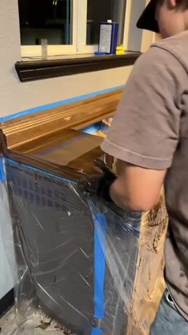Two people are working in a kitchen with dark brown cabinets and a light-colored backsplash. They are applying a clear, glossy finish to a wooden countertop. The countertop has an integrated sink cutout. The surrounding areas, including the floor and cabinets, are covered with blue painter's tape and clear plastic sheeting for protection. One person, wearing black gloves, is using a squeegee to spread the finish. The other person, wearing a baseball cap and a t-shirt, is also working on the countertop. Several containers and tools are visible on the floor.