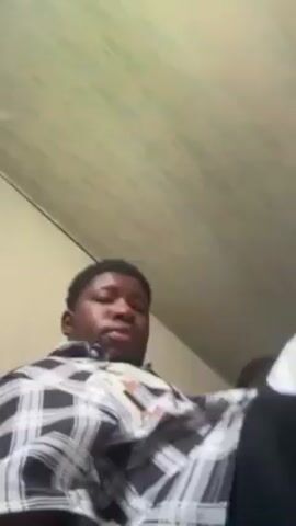 The video shows a room filled with seated people. The camera is held by one of the people in the room, who appears to be facing forward. The room has a low ceiling with at least one ceiling fan. Most people in the room are dark-skinned. Many people are wearing casual clothing, including shirts with stripes or geometric patterns. The camera moves around the room, briefly showing the faces of several individuals and the arrangement of the seats. The lighting is somewhat dim, and the video is slightly shaky.