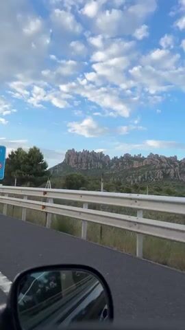 The video shows a view from a car driving along a road. In the foreground, the side mirror of the car is visible. Ahead, there is a highway with a white metal guardrail. A blue road sign indicating a tunnel named "Túnel del Bruc" is visible, along with traffic restriction symbols and route information. Beyond the road and guardrail, there are green trees and a mountainous landscape with distinct rock formations under a blue sky with scattered clouds. The camera pans slightly, capturing the passing scenery.
